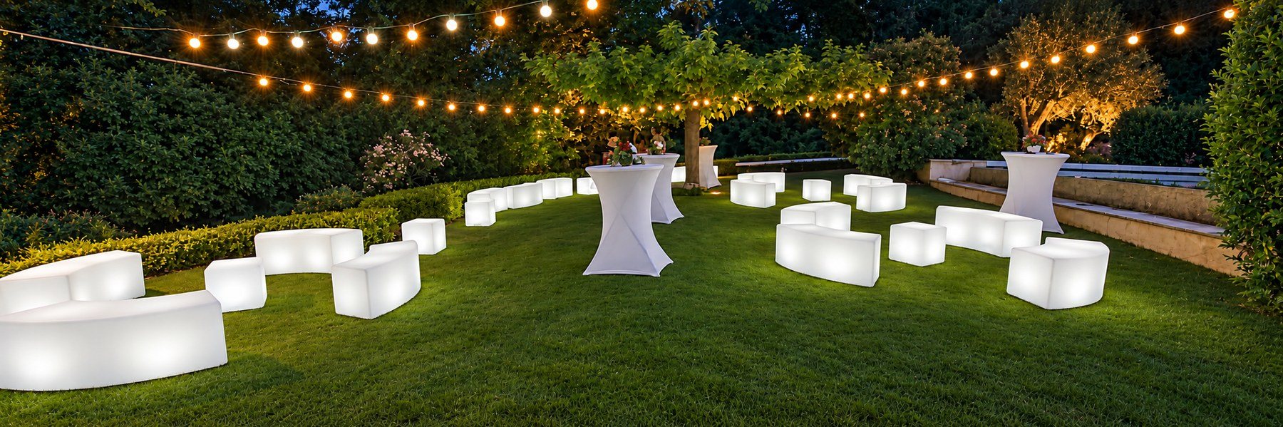 Bancs lumineux blancs et cubes disposés sur une pelouse sous des guirlandes de nuit